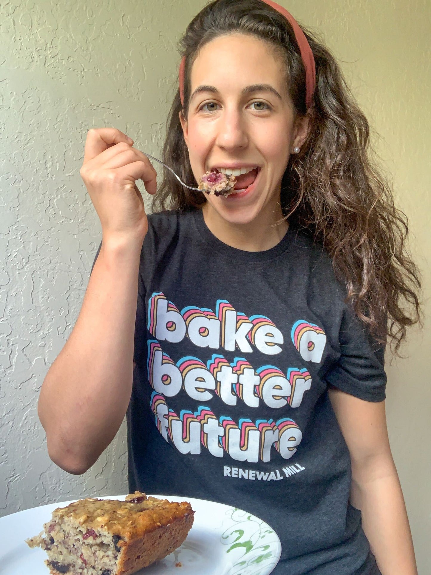 Woman eating a baked good wearing a charcoal heather unisex t-shirt with 'Bake a Better Future' retro slogan made from 100% recycled fabric